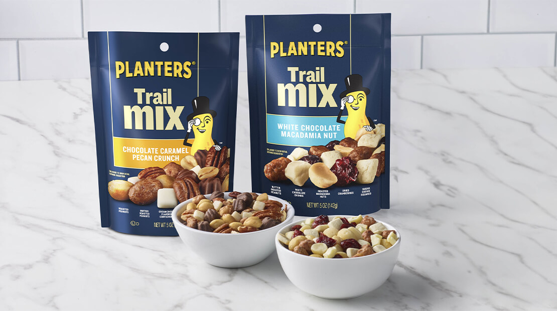 Two new varieties of PLANTERS® Trail Mix sitting on a marble countertop. In front of the two bags are small bowls of each trail mix variety.
