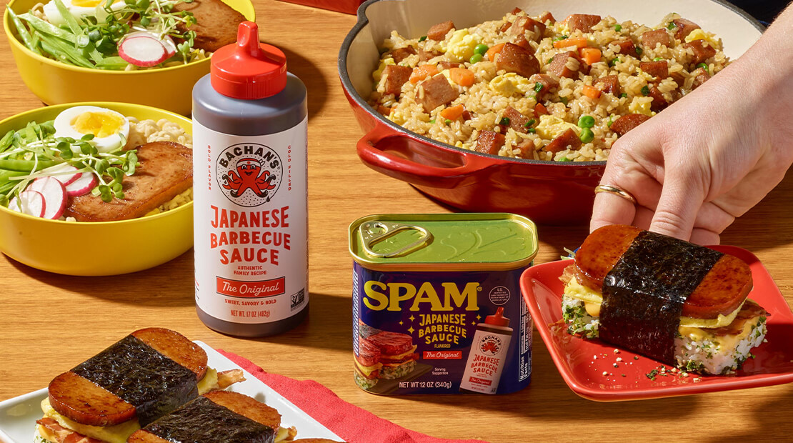 An assortment of dishes made with SPAM® products and Bachan’s Japanese Barbecue Sauce displayed on a wooden table. A large bowl of SPAM® fried rice sits in the background, two bowls of noodle soup with sliced radishes and greens are on the left, and several SPAM® musubi pieces wrapped in nori are arranged on plates. A hand is placing one musubi on a red plate. Bottles and cans of SPAM® product and Bachan’s Japanese Barbecue Sauce are positioned in the center.