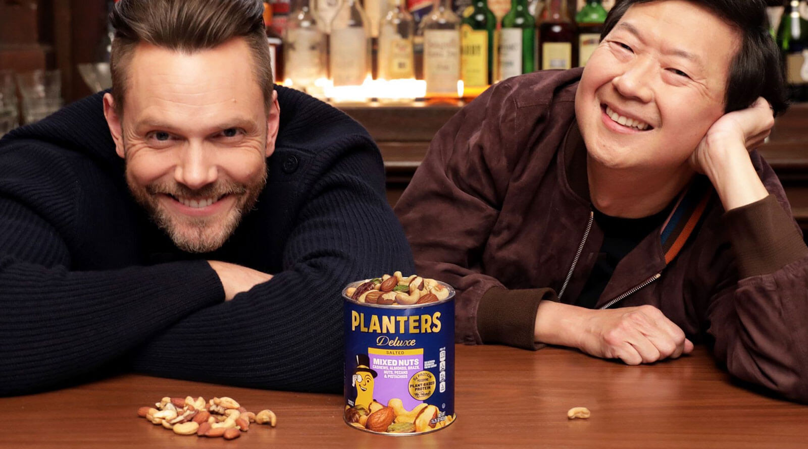 Joel McHale and Ken Jeong leaning on a wooden table with a can of Planters Mixed Nuts on it