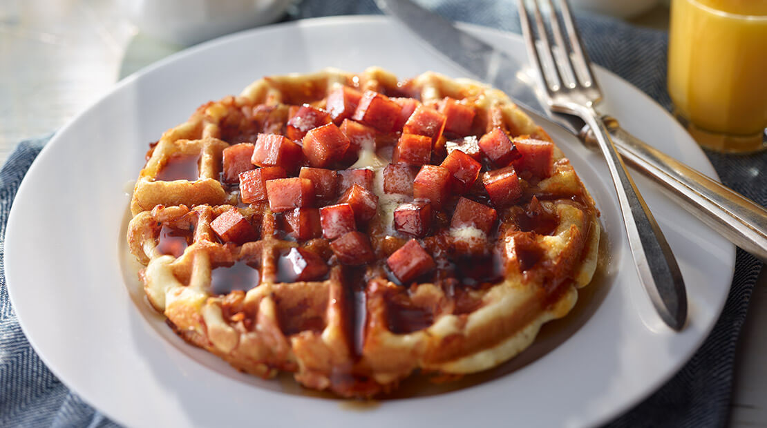 A waffle on a white plate topped with cubed SPAM and maple syrup