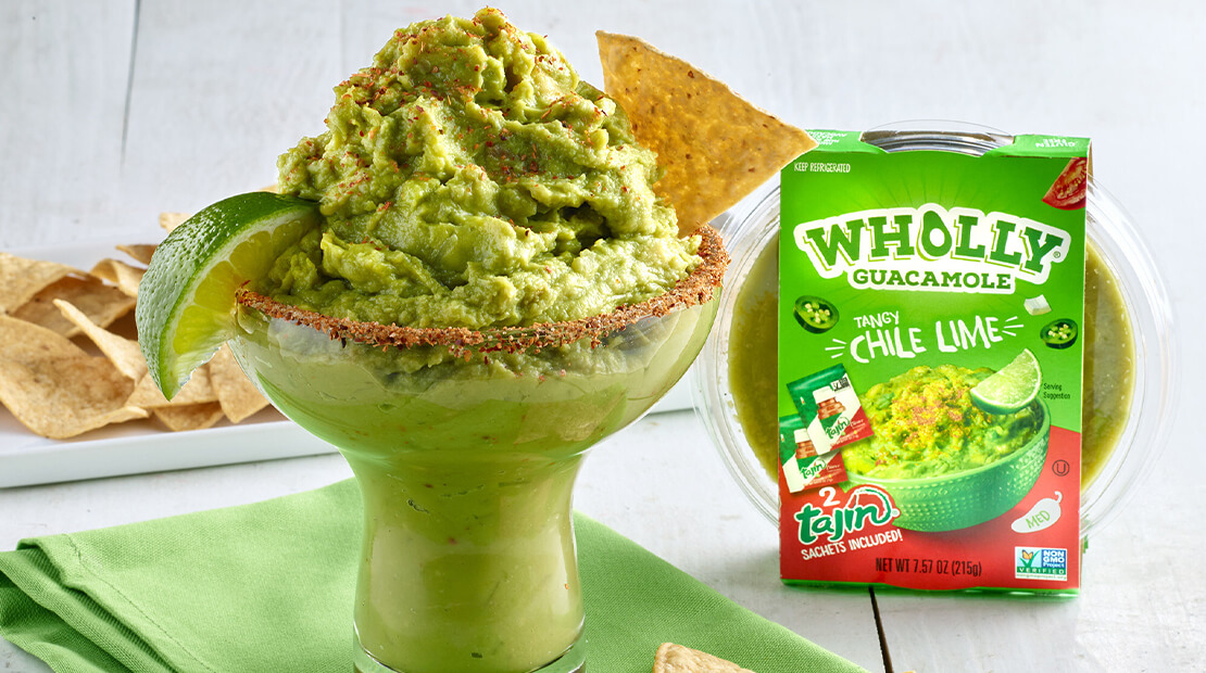 WHOLLY® GUACAMOLE Chile Lime in a large glass with a wedge of lime on the rim and a tortilla chip dipped into the guacamole.