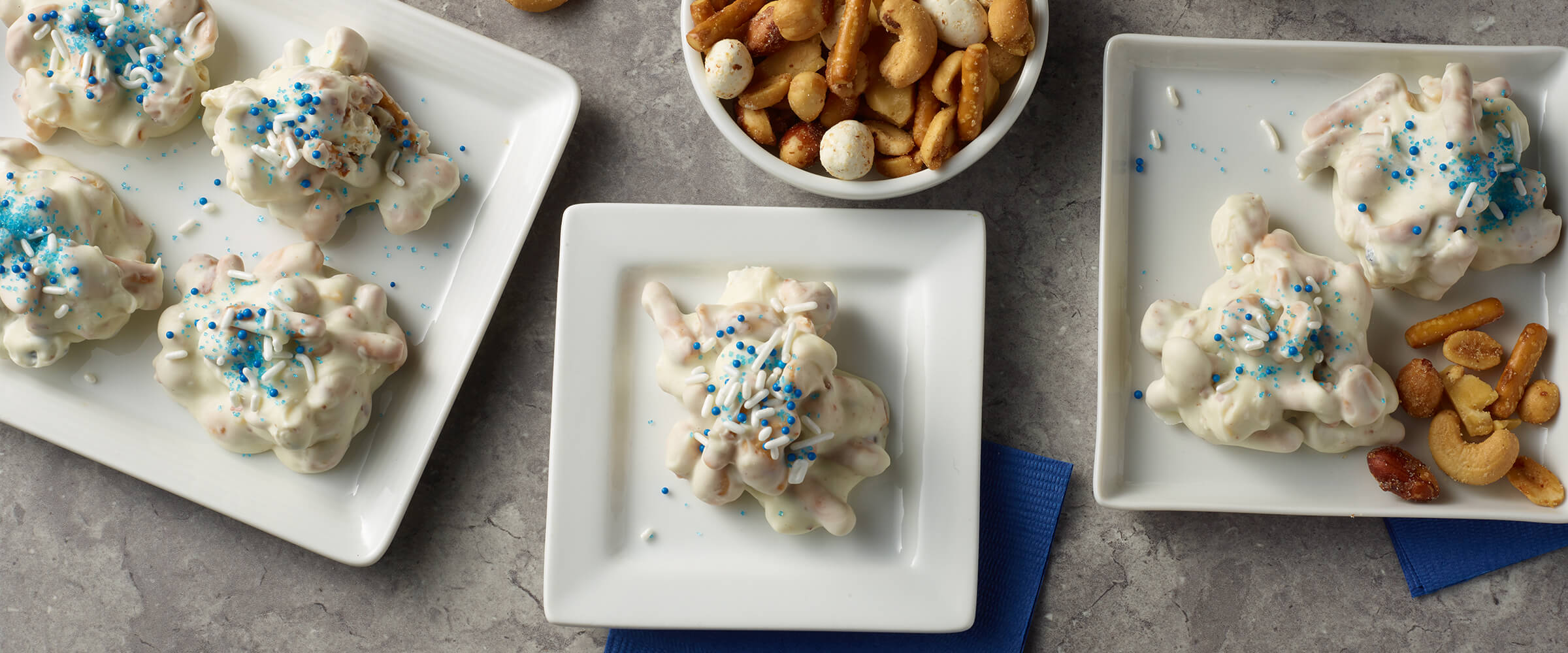 7 Brittle Nut Medley Candy Clusters on three small white square plates, Next to the plates in a small round dish of Brittle Nut Medley.