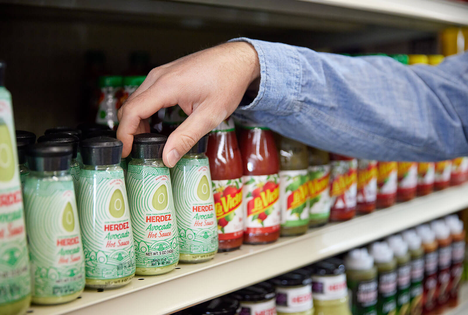 A hand reaching out and grabbing a bottle of Herdez Avocado hot sauce